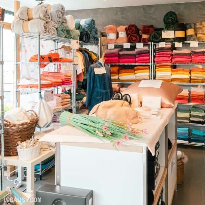 L'intérieur du magasin de linge de maison Comptoir du Linge à Beaufays, avec des étagères métalliques remplies de serviettes et de draps de différentes couleurs, soigneusement pliés et empilés. Il y a également des peignoirs suspendus et divers articles de literie exposés sur une table centrale, y compris un oreiller, des chaussons et des fleurs décoratives.