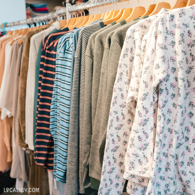 Une rangée de vêtements suspendus sur des cintres en bois dans le magasin de linge de maison Comptoir du Linge à Beaufays. Les vêtements incluent des chemises à rayures, des pulls en tricot, et des chemises à motifs floraux.