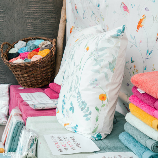 Un panier en osier rempli de serviettes de différentes couleurs, des piles de serviettes pliées de couleurs vives (rose, orange, jaune, bleu, gris), et un coussin avec un motif floral dans le magasin de linge de maison Comptoir du Linge à Beaufays. Il y a également des étiquettes avec des informations sur les produits.