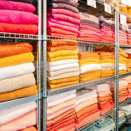 Des étagères métalliques remplies de serviettes de différentes couleurs dans magasin de linge de maison Comptoir du Linge à Beaufays. Les serviettes sont soigneusement pliées et empilées par couleur, allant du rose au beige. Des étiquettes de prix sont visibles sur certaines étagères, indiquant des prix tels que 2€, 2.5€, et 12€.