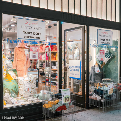 Devanture du magasin de linge de maison Comptoir du Linge à Liège. La vitrine est décorée avec divers articles de linge de maison, tels que des peignoirs, des serviettes et des coussins. Des paniers à l'extérieur contiennent des articles en promotion avec des étiquettes de prix.