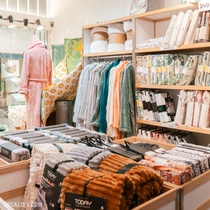 L'intérieur du magasin de linge de maison Comptoir du Linge à Liège, avec des étagères et des présentoirs remplis de divers articles de linge de maison, tels que des peignoirs, des couvertures, des draps et des housses de couette. Un mannequin porte un peignoir rose, ajoutant une touche visuelle intéressante à l'ensemble.