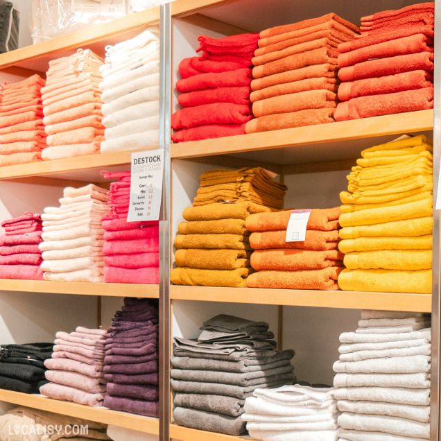 Des étagères remplies de serviettes de différentes couleurs dans le magasin de linge de maison Comptoir du Linge à Liège. Les serviettes sont soigneusement empilées et organisées par couleur, allant du blanc, rose, orange, jaune, gris, au noir. Il y a une pancarte au centre indiquant DESTOCK avec des prix pour différents articles.