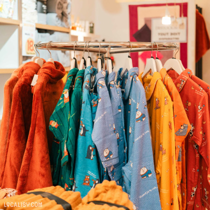 Un présentoir de vêtements dans le magasin de linge de maison Comptoir du Linge à Liège. Sur le présentoir, il y a une variété de pyjamas colorés avec des motifs de personnages de dessins animés. Les pyjamas sont suspendus sur des cintres et sont de différentes couleurs, notamment vert, bleu, jaune et rouge.