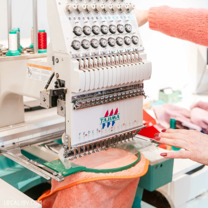 Une machine à broder industrielle de la marque Tajima en train de broder un tissu. La machine est équipée de plusieurs aiguilles et bobines de fil de différentes couleurs. Une main ajuste le tissu ou la machine à l'intérieur du magasin de linge de maison Comptoir du Linge à Liège.