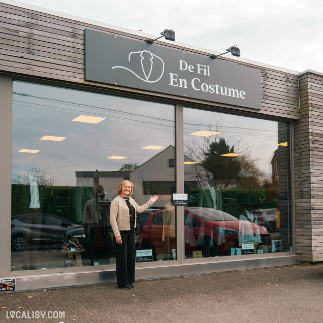 Devanture du magasin de vêtements pour hommes De Fil En Costume à Chaudfontaine. La façade est en bois avec une grande vitrine qui permet de voir l'intérieur du magasin. Une personne se tient devant la vitrine, semblant présenter ou accueillir les visiteurs. Le logo du magasin, représentant un costume stylisé, est visible sur l'enseigne au-dessus de la vitrine.