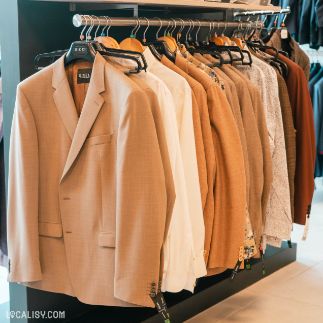 Une rangée de vestes et de chemises suspendues sur des cintres dans le magasin de vêtements pour hommes De Fil En Costume à Chaudfontaine. Les vestes sont principalement de couleur beige et marron, avec quelques chemises blanches et à motifs. Les cintres portent l'étiquette DIGEL.