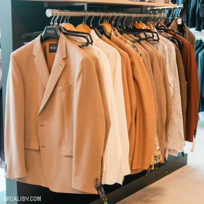 Une rangée de vestes et de chemises suspendues sur des cintres dans le magasin de vêtements pour hommes De Fil En Costume à Chaudfontaine. Les vestes sont principalement de couleur beige et marron, avec quelques chemises blanches et à motifs. Les cintres portent l'étiquette DIGEL.