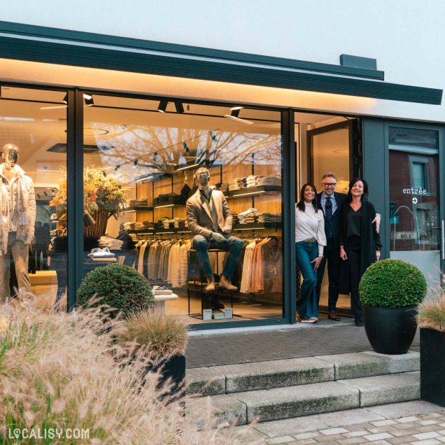 Devanture du magasin de vêtements pour hommes Boutique Pierre Thonon à Beaufays. La vitrine présente des mannequins habillés de manière élégante, avec des vêtements soigneusement disposés sur des étagères à l'intérieur. Devant l'entrée, il y a deux grandes plantes en pot et des marches menant à la porte. Trois personnes se tiennent devant l'entrée, semblant poser pour une photo.