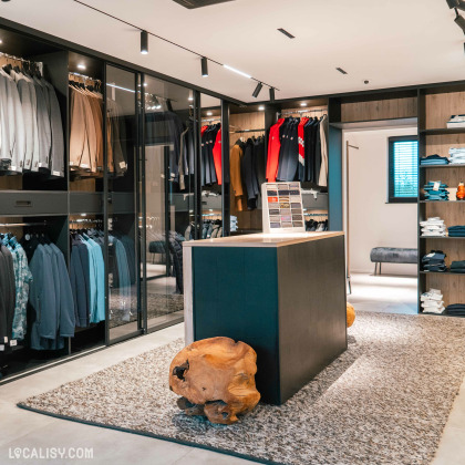 L'intérieur du magasin de vêtements pour hommes Boutique Pierre Thonon à Beaufays. La boutique est bien organisée avec des vêtements soigneusement accrochés et pliés. Il y a des costumes, des vestes et des chemises de différentes couleurs et styles rangés dans des armoires vitrées et sur des étagères.