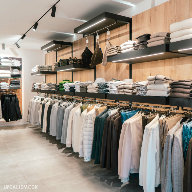 L'intérieur du magasin de vêtements pour hommes Boutique Pierre Thonon à Beaufays, avec des vêtements soigneusement organisés sur des étagères et des cintres. Les étagères contiennent des piles de pulls, de chemises et d'autres vêtements pliés, tandis que les cintres présentent des vestes, des manteaux et des pantalons.