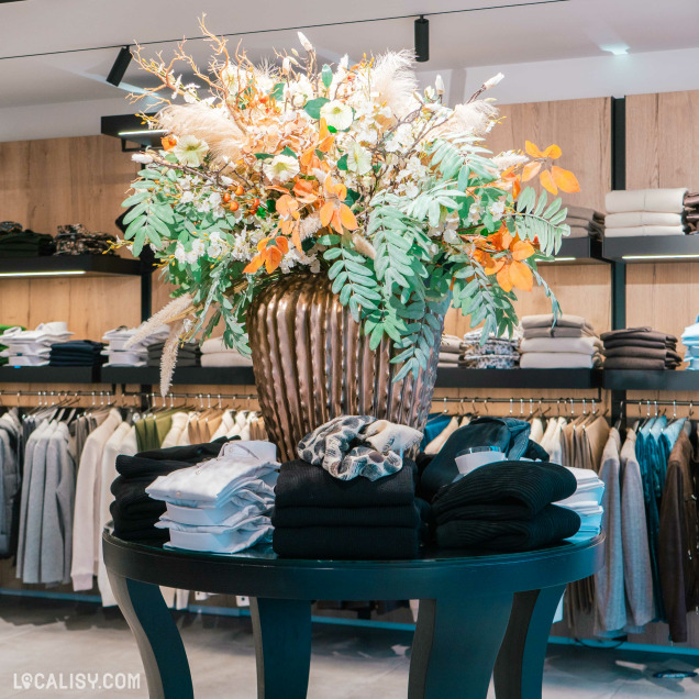 Une grande table ronde noire sur laquelle est posée un grand vase en métal ondulé contenant un arrangement floral luxuriant avec des feuilles vertes et des fleurs aux teintes automnales dans le magasin de vêtements pour hommes Boutique Pierre Thonon à Beaufays. Autour de la table, des piles de vêtements soigneusement pliés, principalement des chemises et des pulls, sont disposées.