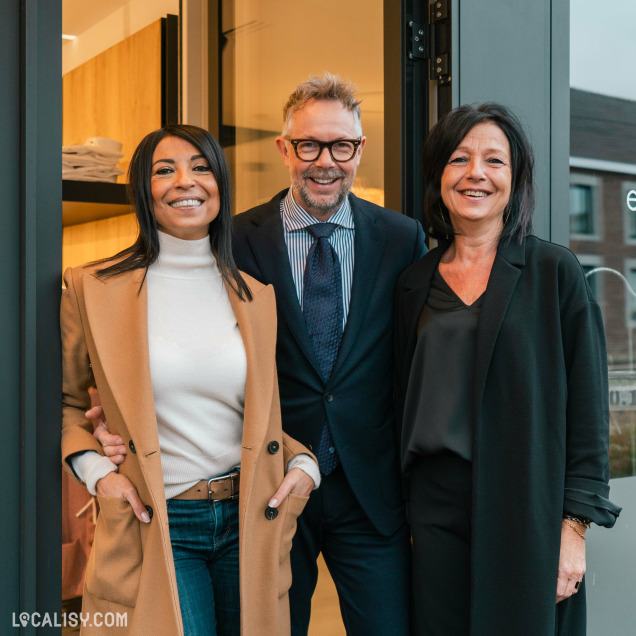 Trois personnes debout devant l'entrée du magasin de vêtements pour hommes Boutique Pierre Thonon à Beaufays. La personne à gauche porte un manteau beige, un pull blanc à col roulé et un jean. La personne au centre porte un costume sombre avec une chemise rayée et une cravate. La personne à droite porte un manteau noir et un haut sombre. L'intérieur du magasin est visible en arrière-plan, avec des étagères contenant des vêtements pliés.