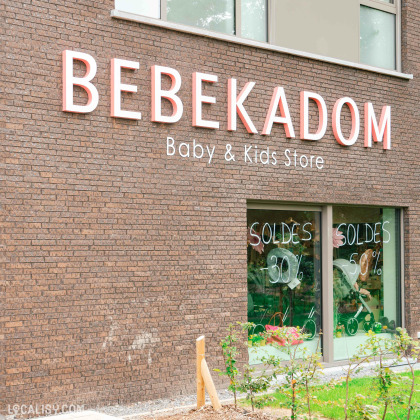 Devanture du magasin de puériculture Bébékadom à Chaudfontaine. La façade en briques brunes arbore une grande enseigne en lettres blanches et roses indiquant BEBEKADOM Baby & Kids Store. Les vitrines affichent des soldes avec des réductions de 30% et 50%, écrites en blanc.