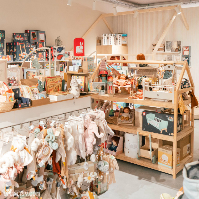 Des étagères en bois remplies de jouets, de peluches, de livres et d'autres articles de puériculture dans le magasin de puériculture Bébékadom à Chaudfontaine. On y voit des vêtements pour bébés suspendus, des jouets éducatifs, des poupées, des accessoires de lit pour bébé, et des jeux de construction.