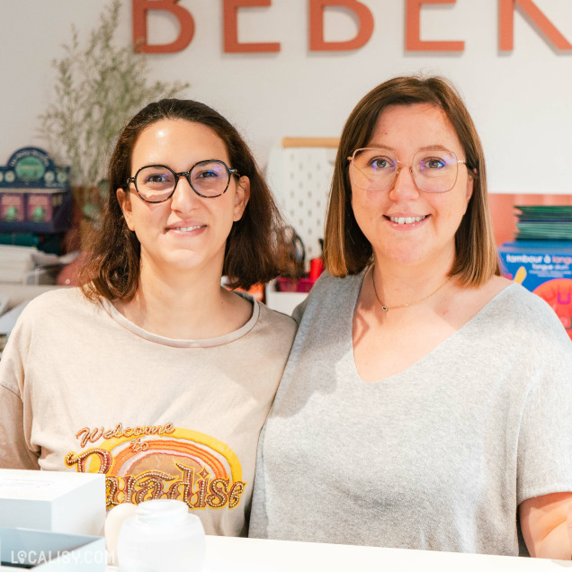 Deux personnes debout à l'intérieur du magasin de puériculture Bébékadom à Chaudfontaine, devant un comptoir. En arrière-plan, on peut voir des étagères avec divers produits pour enfants, ainsi que des décorations colorées. Le nom du magasin "Bébékadom" est visible en grandes lettres rouges sur le mur derrière les personnes.