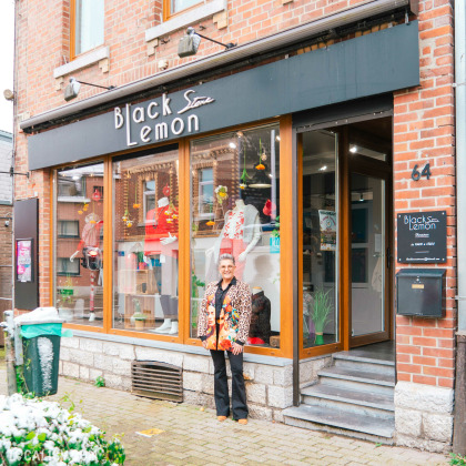 Devanture du magasin de vêtements pour femmes Black Lemon Store à Welkenraedt, avec une façade en briques rouges ornée de grandes vitrines qui exposent des mannequins habillés de vêtements colorés et à la mode. Une enseigne noire avec le nom Black Lemon Store est placée au-dessus des vitrines. À droite de l'entrée, une plaque indique les horaires d'ouverture. Une personne portant une veste colorée se tient devant le magasin.