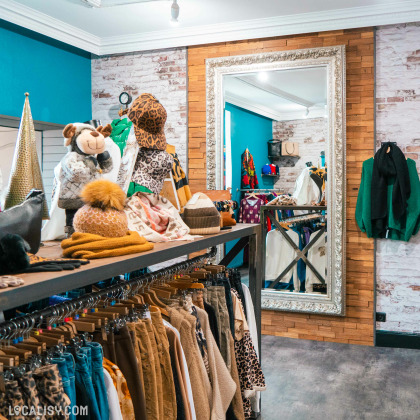 Des vêtements et des accessoires exposés sur des étagères et des portants dans le magasin de vêtements pour femmes Black Lemon Store à Welkenraedt. Il y a un grand miroir encadré de manière décorative sur le mur du fond, entouré de murs en briques et en bois. Des articles comme des chapeaux, des écharpes, des sacs et des vêtements sont soigneusement disposés, créant une ambiance accueillante et organisée.