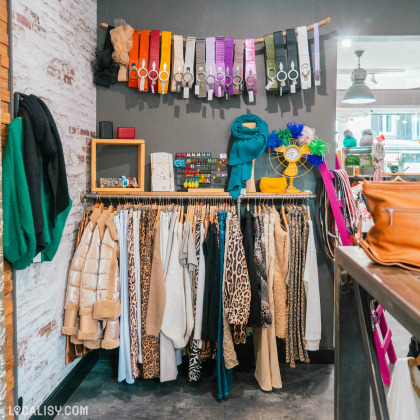 Une variété de vêtements suspendus sur des cintres, incluant des manteaux, des pantalons et des jupes, avec des motifs léopard et des couleurs neutres dans le magasin de vêtements pour femmes Black Lemon Store à Welkenraedt. Au-dessus des vêtements, il y a une rangée de ceintures colorées accrochées à une branche, ainsi que des accessoires comme des écharpes, des montres et des bijoux.