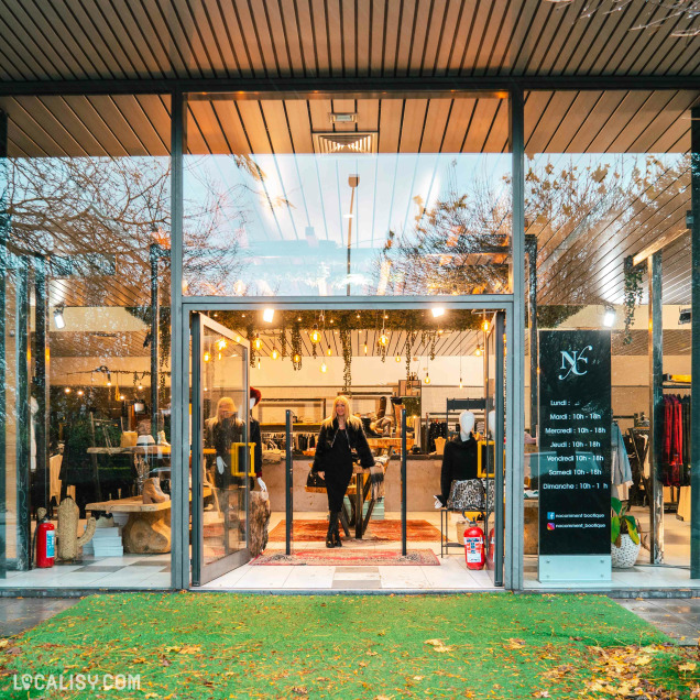 Devanture du magasin de vêtements pour femmes No Comment à Soumagne. La façade est en verre, permettant de voir l'intérieur du magasin où plusieurs mannequins et vêtements sont exposés. À droite de l'entrée, un panneau affiche les horaires d'ouverture du magasin.