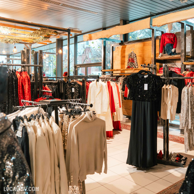 L'intérieur du magasin de vêtements pour femmes No Comment à Soumagne, avec plusieurs portants de vêtements, principalement des robes et des hauts, dans des tons variés incluant le noir, le rouge, le blanc et des paillettes dorées. Le magasin est bien éclairé avec des ampoules suspendues et dispose de grandes fenêtres laissant entrer la lumière naturelle.