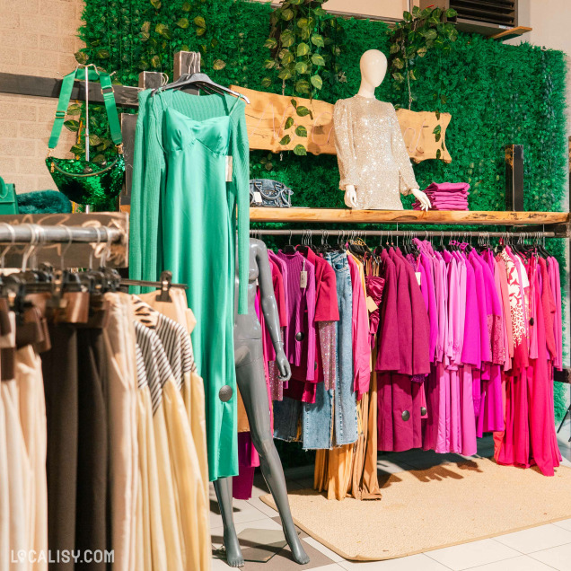 L'intérieur du magasin de vêtements pour femmes No Comment à Soumagne, avec des vêtements suspendus sur des cintres, organisés par couleur. À gauche, il y a une robe verte longue et fluide, et à droite, une rangée de vêtements dans des tons de rose et de violet. Un mannequin porte un haut à paillettes argentées.