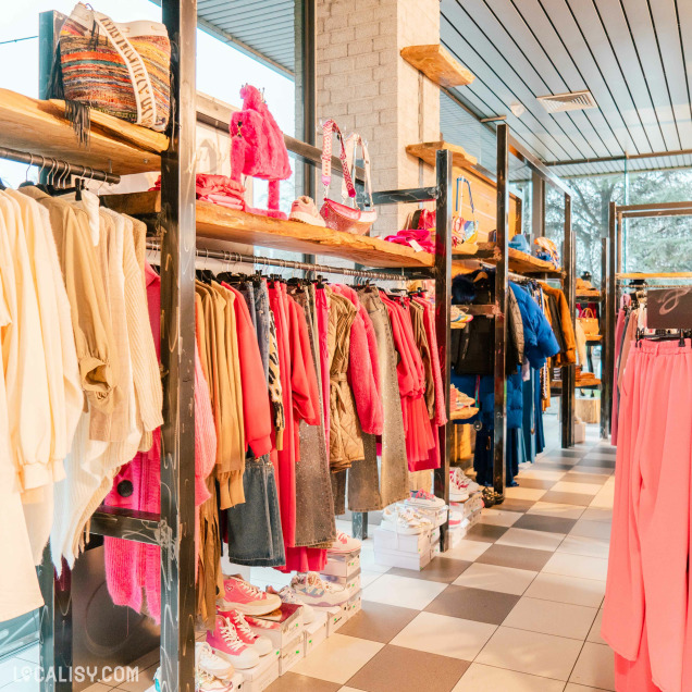 Des vêtements suspendus sur des portants, principalement dans des tons de rose, beige et bleu dans le magasin de vêtements pour femmes No Comment à Soumagne. Il y a également des sacs à main et des chaussures exposés sur des étagères en bois au-dessus des vêtements. Le sol est carrelé avec des carreaux de couleur blanche et grise.