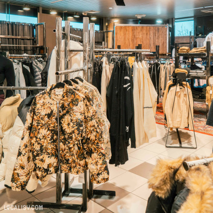 Plusieurs portants de vêtements, principalement des vestes et des manteaux dans le magasin de vêtements pour femmes No Comment à Soumagne. Au premier plan, une veste à motifs floraux est mise en avant. L'agencement du magasin est moderne avec des portants en métal et un sol carrelé.