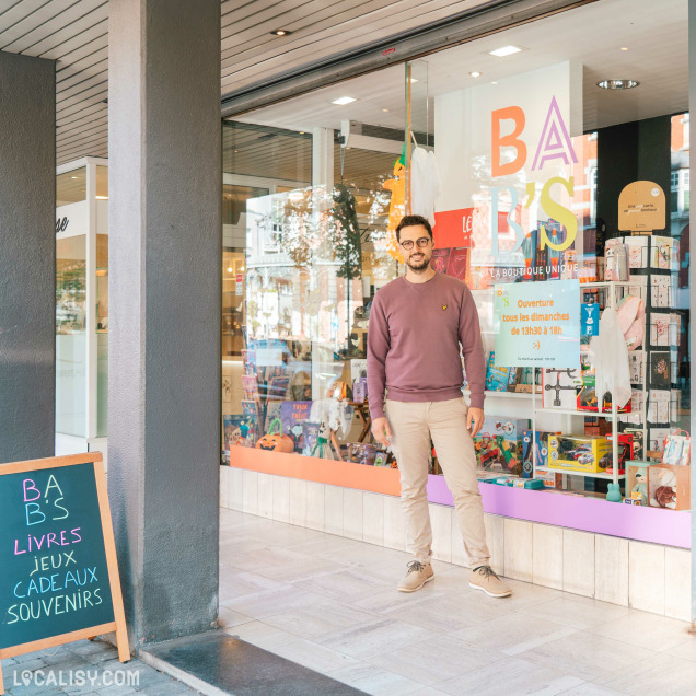 Devanture de la librairie Bab's à Spa. Une personne se tient debout devant la librairie. La vitrine de la librairie est remplie de livres, de jeux, de cadeaux et de souvenirs. Une pancarte à l'extérieur indique BAB'S LIVRES JEUX CADEAUX SOUVENIRS. Une affiche sur la vitrine indique les horaires d'ouverture : Ouverture tous les dimanches de 13h30 à 18h.