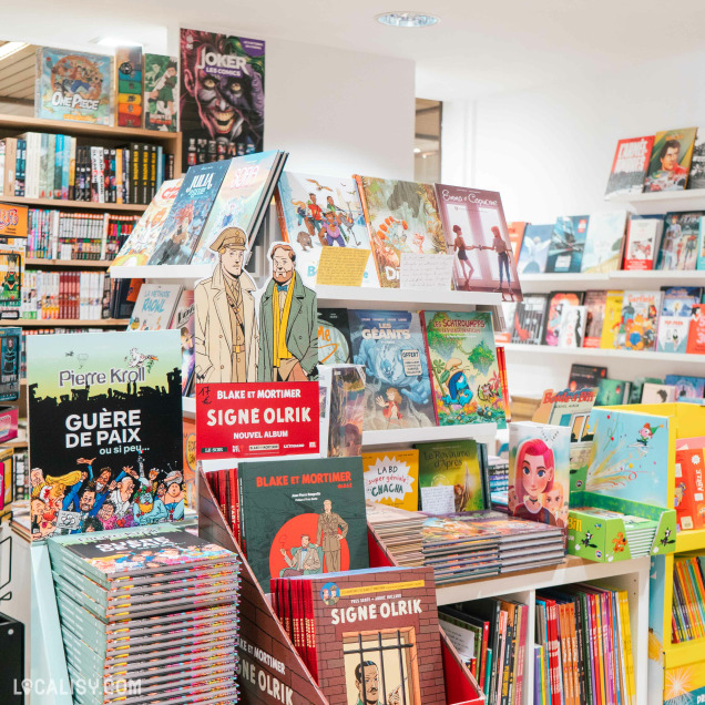 Une section de la librairie Bab's à Spa, avec plusieurs étagères remplies de bandes dessinées et de livres graphiques. Au premier plan, il y a une présentation de livres de la série Blake et Mortimer avec des panneaux publicitaires pour le nouvel album Signe Olrik. On peut également voir des livres de Pierre Kroll intitulés Guerre de Paix ou si peu... et d'autres bandes dessinées populaires comme One Piece et Joker.