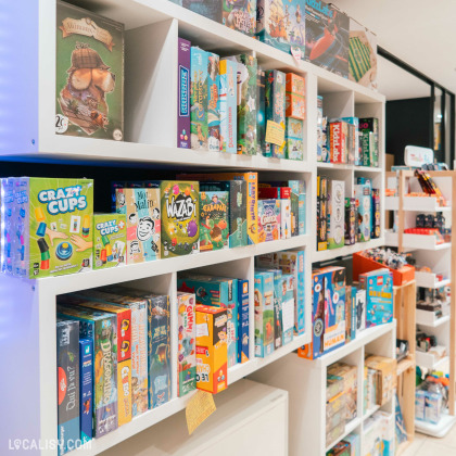 Une étagère remplie de jeux de société dans la librairie Bab's à Spa. Les jeux sont variés et colorés, avec des titres comme Crazy Cups, Wazabi, Carapate, et Mot Malin. L'étagère est bien organisée, avec des jeux empilés et alignés, ce qui donne une impression de diversité et d'abondance.