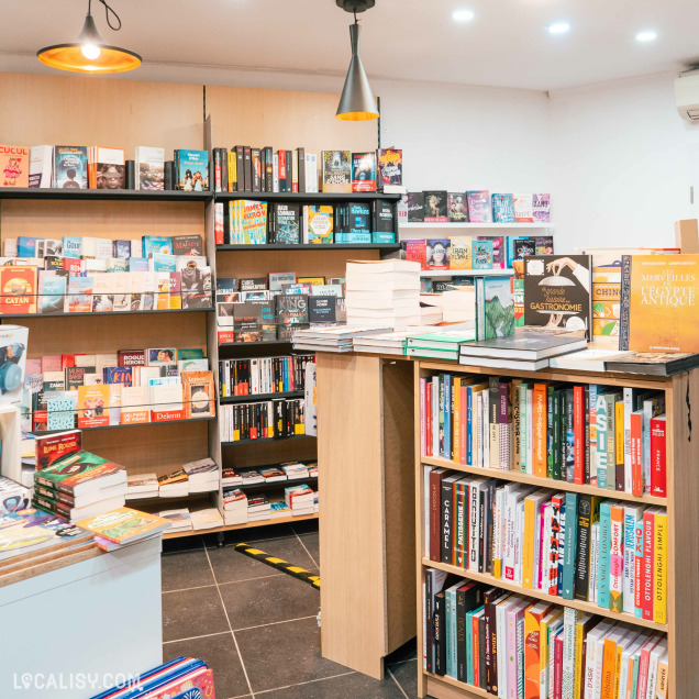 L'intérieur de la librairie Bab's à Spa, avec plusieurs étagères remplies de livres de différents genres et tailles. Il y a des livres empilés sur une table centrale ainsi que sur les étagères. L'éclairage est chaleureux avec des lampes suspendues au plafond.