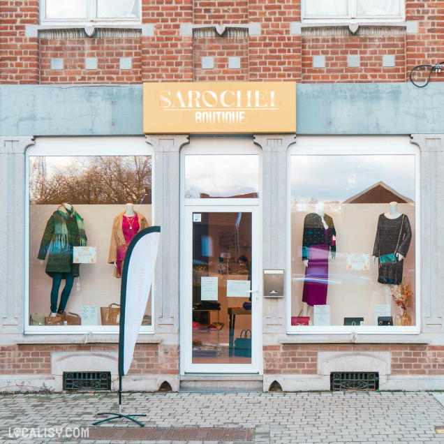 Devanture du magasin de vêtements pour femmes Boutique Sarochel à Flémalle. La façade est en briques rouges avec une enseigne jaune portant le nom SAROCHEL BOUTIQUE. La vitrine expose plusieurs mannequins habillés de différentes tenues, dont des robes et des manteaux. Une banniére publicitaire est placée à gauche de l'entrée principale. La porte d'entrée est en verre, avec des affiches collées dessus.