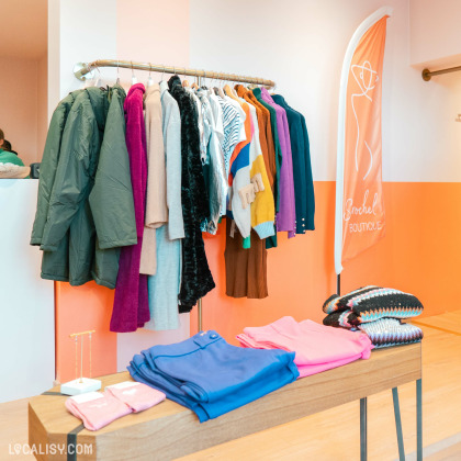 Une variété de vêtements suspendus sur une tringle, y compris des manteaux verts, des pulls de différentes couleurs, et des vêtements à motifs dans le magasin de vêtements pour femmes Boutique Sarochel à Flémalle. Sur une table en bois devant le présentoir, il y a des vêtements pliés, notamment des pantalons bleus et roses, ainsi que des pulls tricotés. Un drapeau avec le logo de la boutique et le texte Sarochel Boutique est visible à droite.