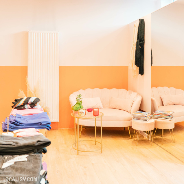 L'intérieur du magasin de vêtements pour femmes Boutique Sarochel à Flémalle. On y voit un coin salon avec un canapé beige, une petite table ronde dorée avec des objets décoratifs, et des piles de magazines. À gauche, il y a une pile de vêtements pliés de différentes couleurs. Le mur est peint en deux tons, orange en bas et blanc en haut, et il y a un grand miroir à droite.