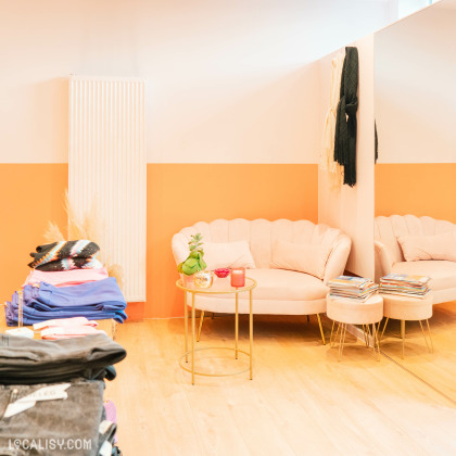 L'intérieur du magasin de vêtements pour femmes Boutique Sarochel à Flémalle. On y voit un coin salon avec un canapé beige, une petite table ronde dorée avec des objets décoratifs, et des piles de magazines. À gauche, il y a une pile de vêtements pliés de différentes couleurs. Le mur est peint en deux tons, orange en bas et blanc en haut, et il y a un grand miroir à droite.