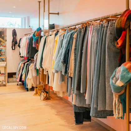 Des vêtements suspendus sur des cintres le long d'une tringle, avec une variété de jeans, de vestes, de chemises et d'autres articles de mode dans le magasin de vêtements pour femmes Boutique Sarochel à Flémalle. À droite, il y a des écharpes colorées accrochées.