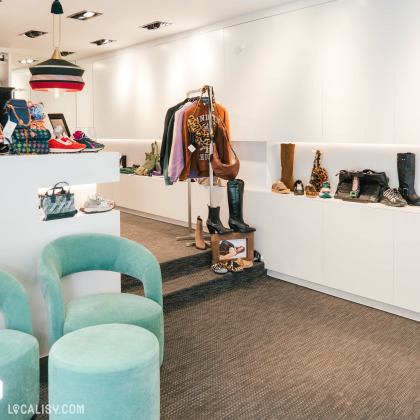 L'intérieur du magasin de vêtements Icon Store à Liège, avec des murs blancs et un sol en moquette grise. À gauche, il y a deux fauteuils verts et un petit tabouret assorti. Sur la gauche, il y a également un présentoir avec des chaussures et des sacs à main. Au centre, un portant expose plusieurs vêtements, dont des vestes et des pulls. À droite, des étagères blanches présentent divers articles, principalement des chaussures et des bottes.