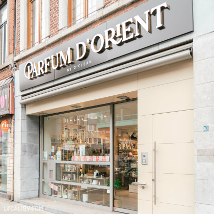 Devanture de la Parfumerie Parfum d'Orient à Liège avec une grande vitrine qui permet de voir l'intérieur du magasin. L'enseigne est en lettres blanches sur un fond noir, avec le sous-titre BY O'CLEAN. On peut également voir des produits exposés à l'intérieur et des affiches de soldes sur la vitrine. Le numéro de rue "63" est visible sur le côté droit de l'entrée.