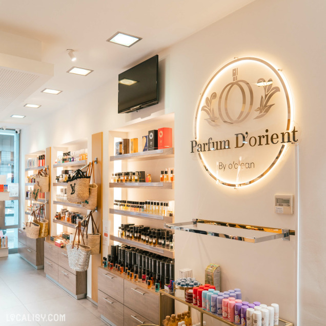 L'intérieur de la Parfumerie Parfum d'Orient à Liège avec des étagères remplies de divers produits de parfumerie, tels que des bouteilles de parfum et des boîtes de produits de beauté. Il y a également des paniers en osier accrochés sur les côtés des étagères. Un grand logo lumineux de la parfumerie est visible sur le mur à droite.