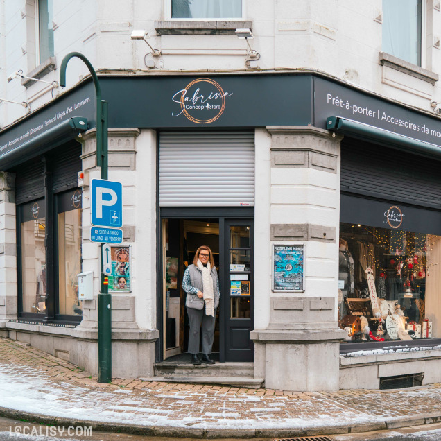 Devanture du magasin de décoration d’intérieur Sabrina Concept Store à Herve. Le magasin est situé à un coin de rue pavée, avec une enseigne noire portant le nom du magasin en lettres cursives blanches et dorées. À gauche de l'entrée principale, il y a une vitrine exposant divers articles de décoration. Un panneau de stationnement bleu est visible à gauche.