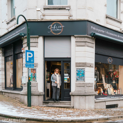 Devanture du magasin de décoration d’intérieur Sabrina Concept Store à Herve. Le magasin est situé à un coin de rue pavée, avec une enseigne noire portant le nom du magasin en lettres cursives blanches et dorées. À gauche de l'entrée principale, il y a une vitrine exposant divers articles de décoration. Un panneau de stationnement bleu est visible à gauche.