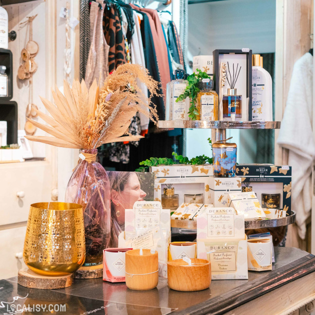 Un présentoir dans le magasin de décoration d’intérieur Sabrina Concept Store à Herve, contenant divers articles de décoration tels que des bougies, des diffuseurs de parfum, des plantes artificielles, et des vases. Les produits sont disposés de manière esthétique sur plusieurs niveaux, avec des étiquettes de prix visibles sur certains articles. En arrière-plan, on peut voir des vêtements suspendus, suggérant que le magasin vend également des articles de mode.