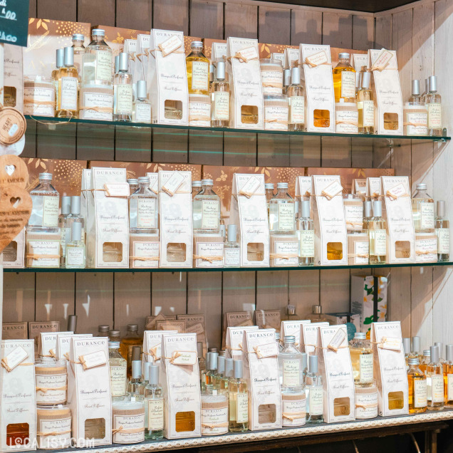 Des étagères en verre dans le magasin de décoration d’intérieur Sabrina Concept Store à Herve, remplies de divers produits de la marque Durance. Les produits incluent des parfums d'ambiance, des diffuseurs de parfum et des bougies, tous présentés dans des emballages élégants et uniformes. Les étiquettes des produits sont principalement blanches avec des accents dorés et des illustrations délicates.