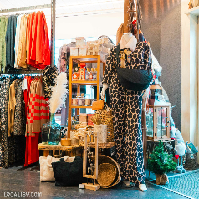 Un mannequin vêtu d'une tenue à motifs de girafe, portant un sac noir en bandoulière dans le magasin de décoration d’intérieur Sabrina Concept Store à Herve. Autour du mannequin, il y a divers articles de décoration et de mode, y compris des vêtements suspendus sur des cintres à gauche, des étagères avec des produits de beauté et des accessoires, ainsi que des objets décoratifs comme des paniers, des coussins, et des plantes.