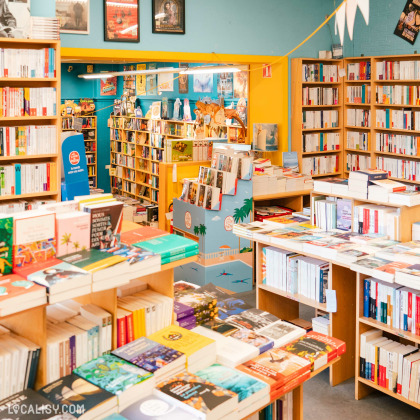 L'intérieur de la librairie L’Oiseau Lire à Visé, avec plusieurs étagères remplies de livres, ainsi que des tables couvertes de livres disposés de manière organisée. Les murs sont peints en bleu et jaune, et il y a des affiches et des décorations colorées accrochées.