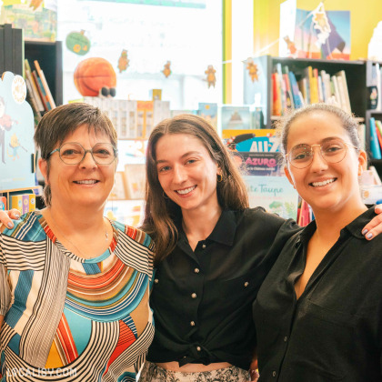 Trois personnes debout dans la librairie L’Oiseau Lire à Visé. En arrière-plan, on peut voir des étagères remplies de livres et des décorations colorées. La librairie semble être bien organisée et accueillante, avec une variété de livres exposés. Il y a également des affiches et des panneaux indiquant différentes sections de la librairie.