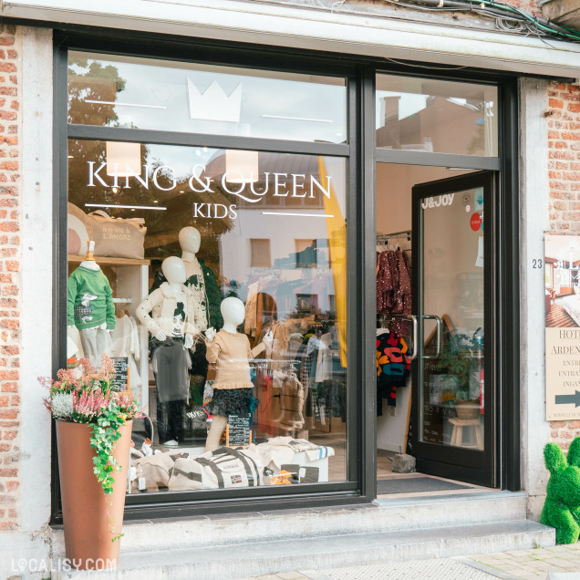 Devanture du magasin de vêtements pour enfants et bébés King & Queen Kids à Spa. La vitrine est en verre avec le nom du magasin écrit en lettres blanches, accompagné d'une couronne stylisée au-dessus. À l'intérieur, on peut voir plusieurs mannequins habillés avec des vêtements pour enfants, ainsi que divers articles de mode exposés. À l'extérieur, il y a un grand pot de fleurs avec des plantes.