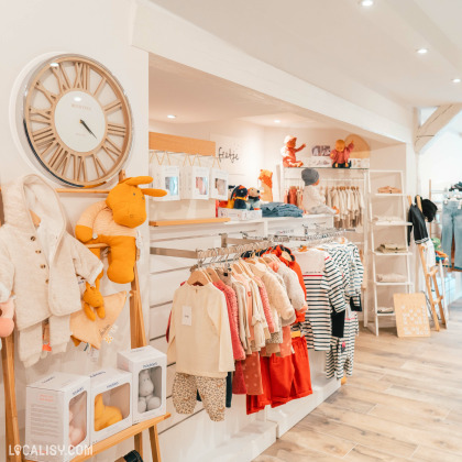 L'intérieur du magasin de vêtements pour enfants et bébés King & Queen Kids à Spa. On y voit une variété de vêtements pour enfants soigneusement exposés sur des étagères et des portants. Il y a également des peluches et des articles emballés en boîte. Le magasin a un agencement chaleureux et bien organisé avec un sol en bois et un éclairage lumineux.