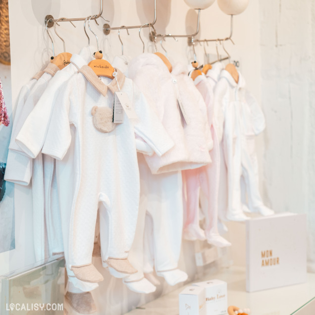 Plusieurs combinaisons et pyjamas pour bébés sont suspendus sur des cintres en bois, accrochés à une barre métallique dans le magasin de vêtements pour enfants et bébés King & Queen Kids à Spa. Les vêtements sont principalement de couleur blanche et rose pâle, avec des détails doux et pelucheux, certains ayant des motifs d'ours en peluche.